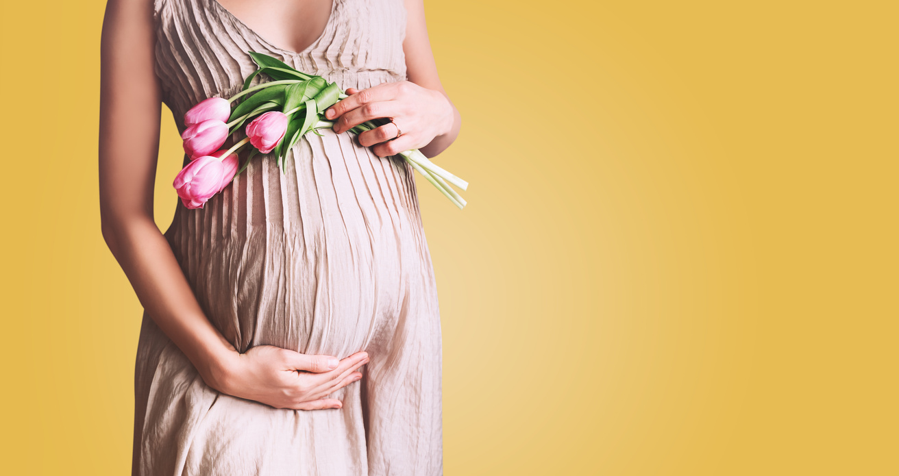 Beautiful pregnant woman with tulips flowers holds hands on belly in white background. Young woman in maternity dress waiting for baby birth. Pregnancy, Motherhood, Mother's Day Holiday concept.